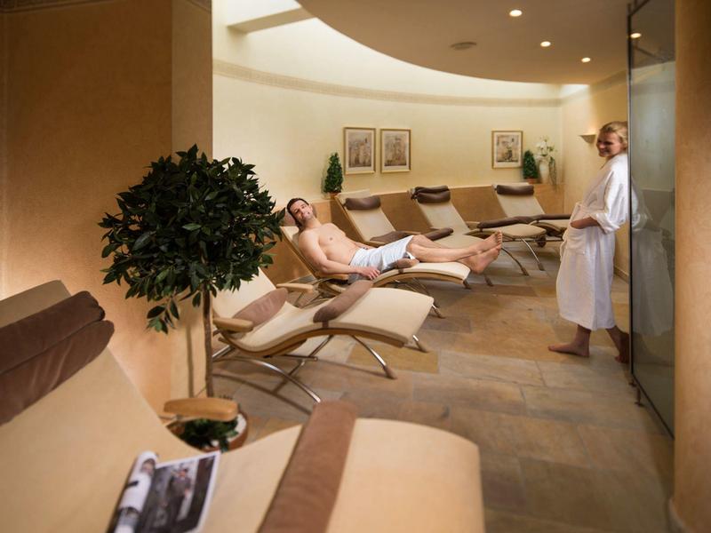 Entspannungsraum mit Liegen, Pflanzen und gedämpftem Licht; zwei Personen in Bademänteln.