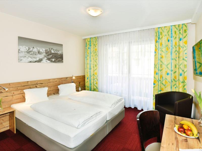 Helles Hotelzimmer mit Doppelbett, Holzakzenten, bunten Vorhängen und Balkonfenster.