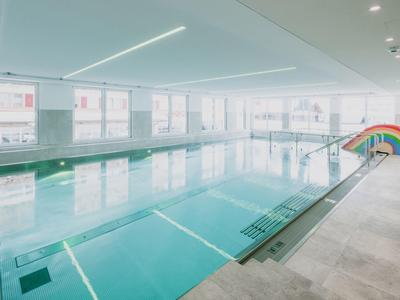 Großes Hallenbad mit klar blauem Wasser, Fenstern und Regenbogen-Rutsche am Rand.