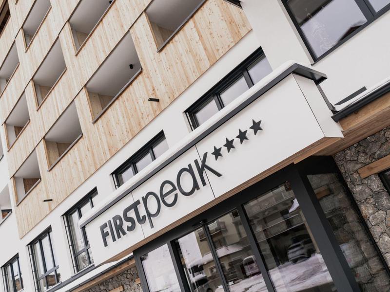 Moderner Hotelbau mit Balkonen, Glasfassade und großem Schriftzug "FIRSTpeak****" am Eingang.