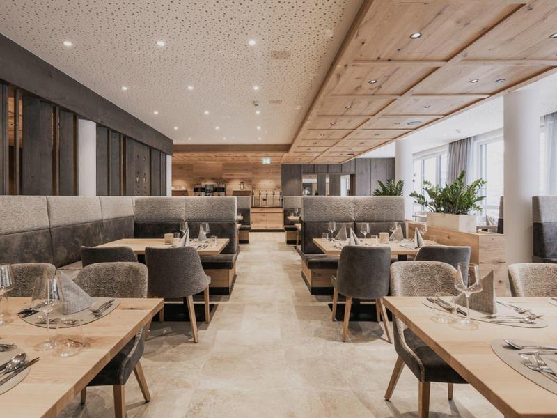 Moderner, heller Restaurantinnenraum mit Holzmöbeln, grauen Sesseln und großen Fenstern.