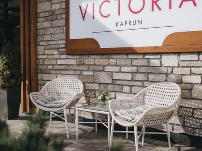 Twee witte stoelen met een klein tafeltje voor Hotel Victoria Kaprun bord aan steenmuur.