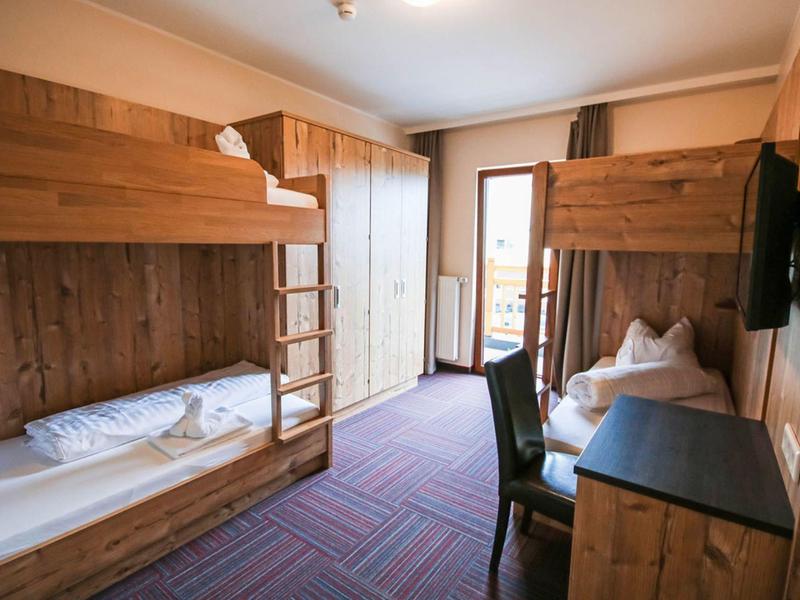Kleines Zimmer mit Holz-Etagenbetten, Teppichboden, Schrank, Stuhl und Balkon mit Tageslicht.