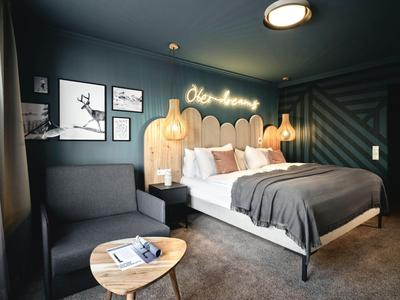 Nowoczesny pokój hotelowy z dużym łóżkiem, szarym fotelem i unikalnym drewnianym zagłówkiem.