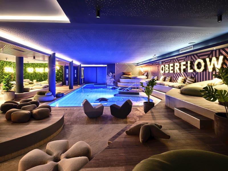 Moderner Indoor-Pool mit Lounge, blauen Lichtern und komfortablen Sitzmöbeln in Holz- und Grau-Tönen.