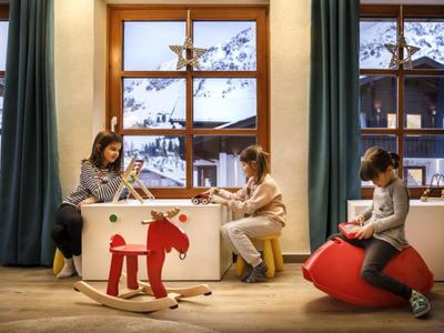 Bambini giocano dentro vicino a una finestra con vista sulla neve, giocattoli in legno e palla rossa.