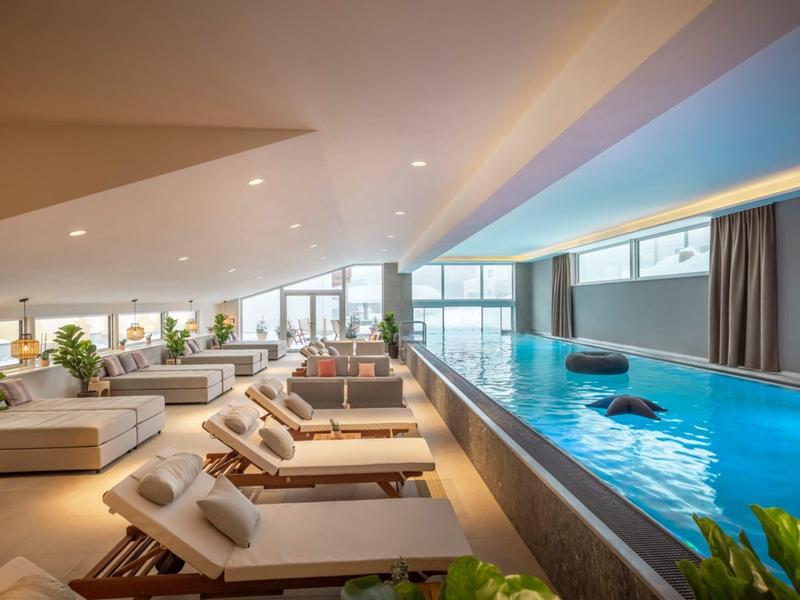 Piscina interna moderna con lettini e grandi finestre luminose.