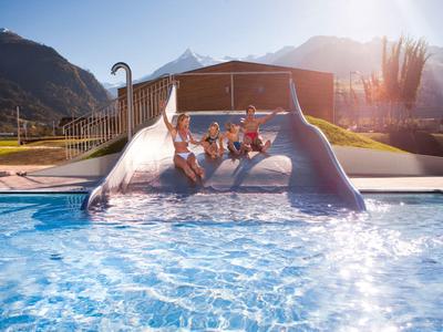 Quattro bambini scendono da uno scivolo ampio in una piscina con montagne sullo sfondo al tramonto.