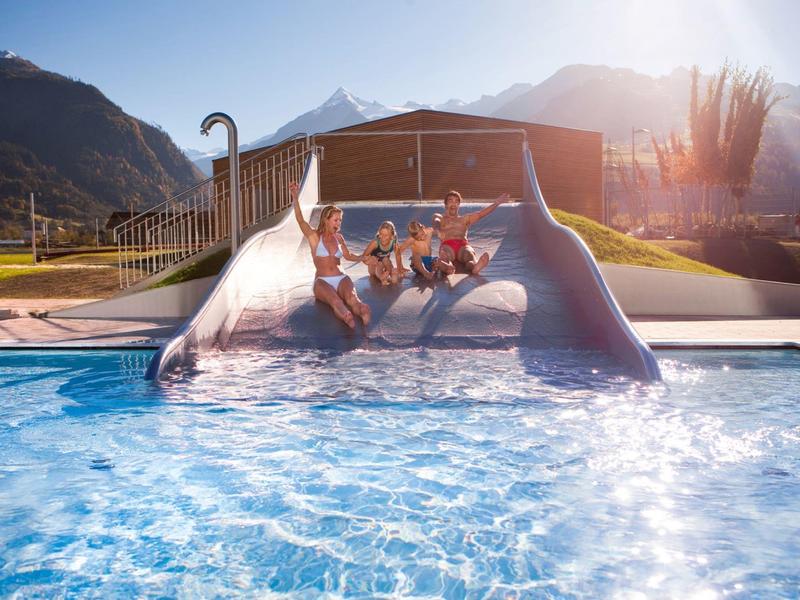 Quattro bambini scendono da uno scivolo ampio in una piscina con montagne sullo sfondo al tramonto.