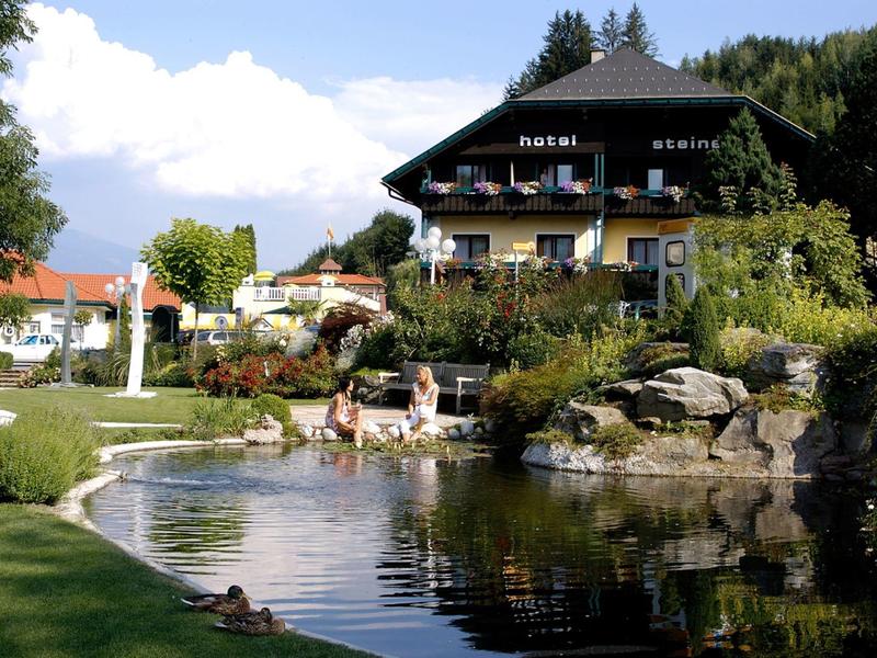 Hotelová budova s balkony vedle rybníka a upravené zahrady v zeleni.