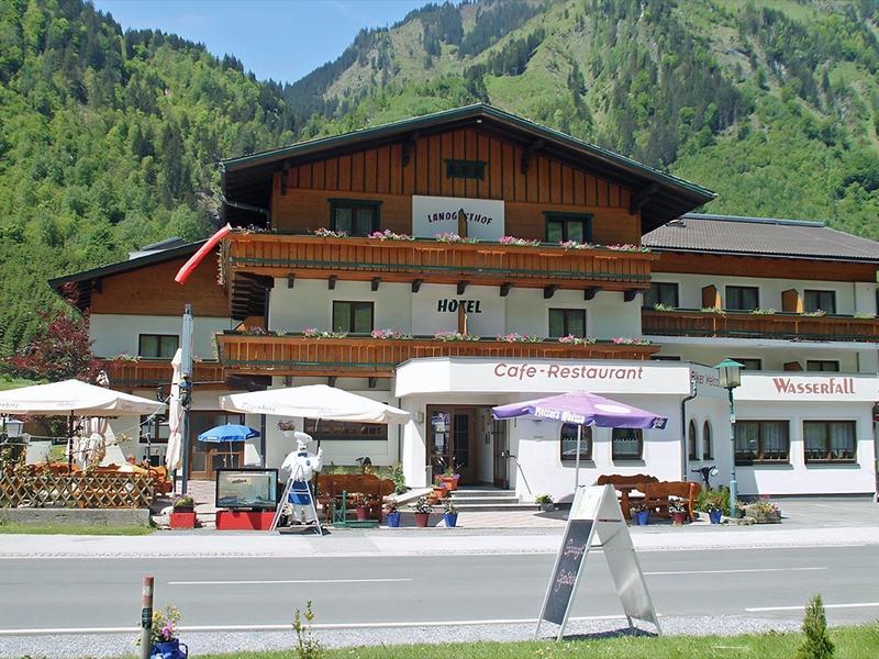 Chalet-style hotel with outdoor seating, umbrellas, and mountain backdrop under clear sky.