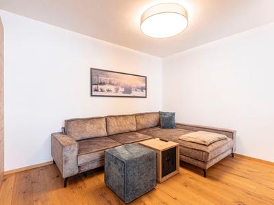 Nowoczesny salon z szarą narożną sofą, dwoma pufami i drewnianą podłogą.