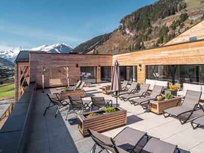 Modernes Berghotel mit sonniger Terrasse und Liegestühlen, umgeben von Bergen und klarem Himmel.