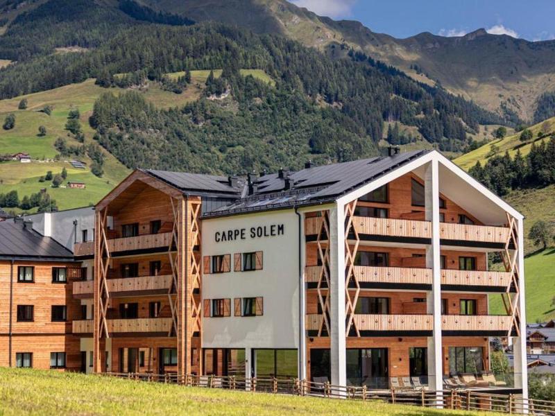 Nowoczesny budynek hotelowy w alpejskim krajobrazie z zielonymi łąkami i górami w tle.