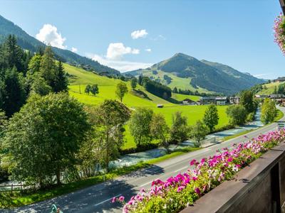 Blick von einem Balkon mit Blumen auf eine Straße und grüne Berge im Sonnenschein.
