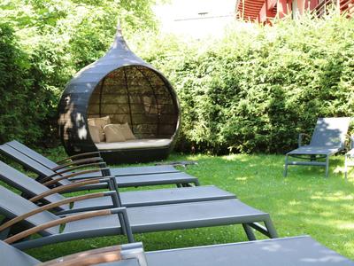 Mehrere Liegestühle und eine runde Hängematte stehen auf einem grünen Rasen vor einem dichten grünen Busch.