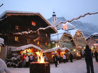 Zimní trh se osvětlenými stánky a otevřeným ohněm před zasněženými dřevěnými domy.