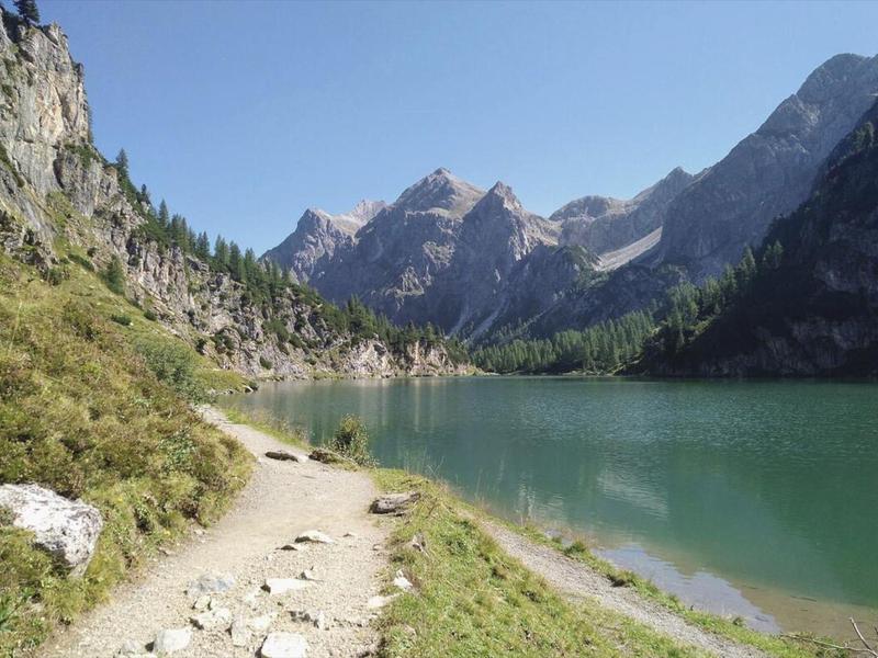 Turistická trasa podél horského jezera s čistou tyrkysovou vodou a skalnatými horami v pozadí