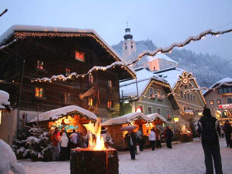 Zimní trh se osvětlenými stánky a otevřeným ohněm před zasněženými dřevěnými domy.