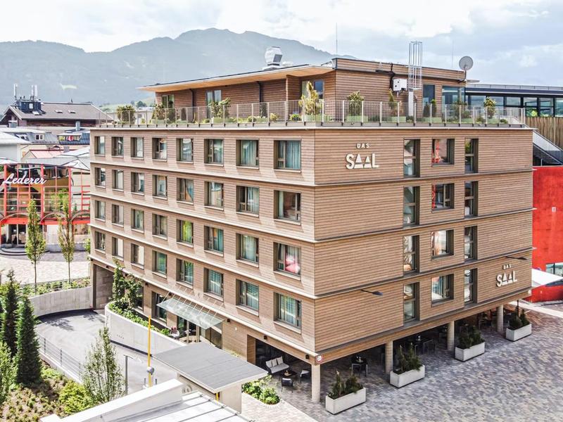 Modernes sechsstöckiges Holzgebäude mit Balkonen und Dachterrasse vor Bergen und bewölktem Himmel.