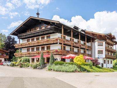 Grande hotel chalet con dettagli in legno, terrazza solare e sfondo montano sotto un cielo limpido.