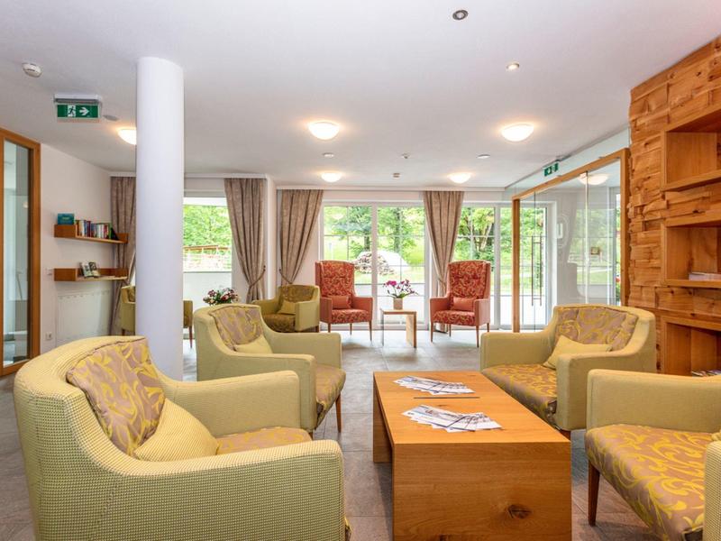 Pohodlná společenská místnost se zelenými křesly a dřevěným stolem v jasné místnosti s velkými okny.