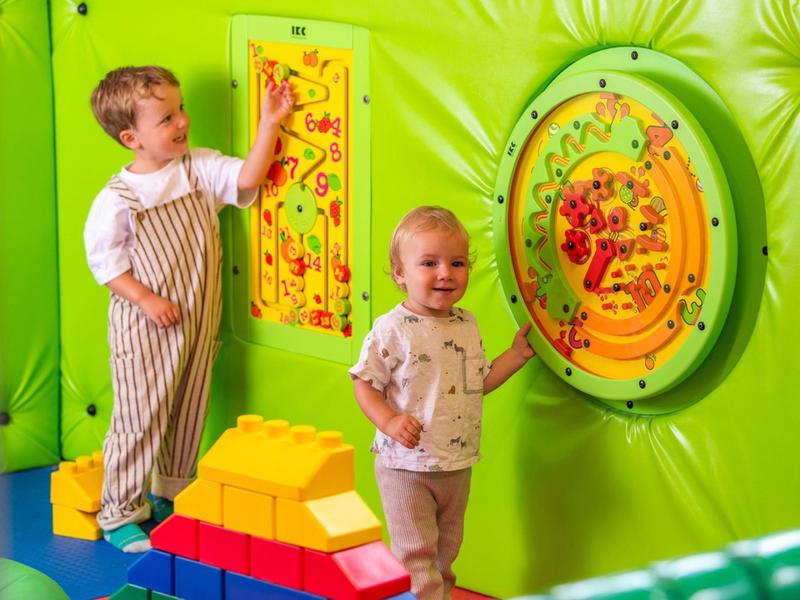 Dvě děti si hrají s barevnými interaktivními hrami na stěně v jasně zelené vnitřní herně.