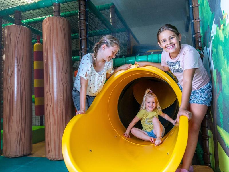 Děti si radostně hrají na žluté skluzavce v kryté herně.