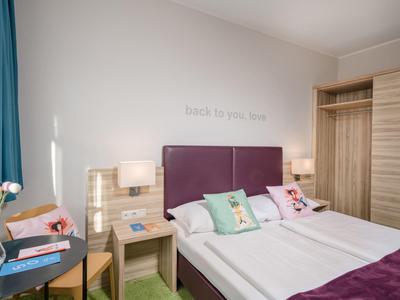 Camera d'albergo luminosa con letto matrimoniale, testiera viola e armadio in legno.