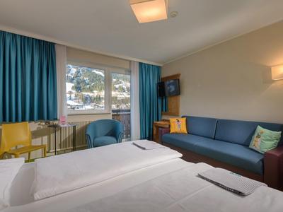 Camera d'hotel con due letti, divano blu, scrivania e poltrona blu vicino alla finestra con tende.