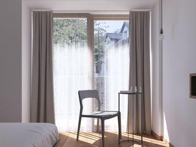 Stanza luminosa e minimalista con finestra, sedia e piccolo tavolo rotondo accanto al letto.