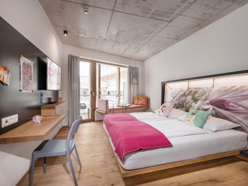 Camera d'hotel moderna con letto grande, scrivania, poltrona e balcone con porte in vetro.