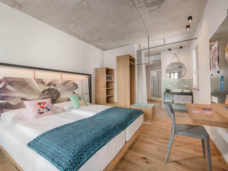 Camera d'hotel arredata modernamente con letto matrimoniale, pavimento in legno e area lavoro