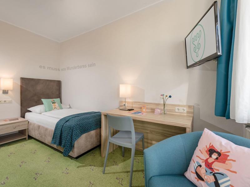 Camera d'hotel accogliente con letto, scrivania e poltrona blu vicino a una finestra con tende.