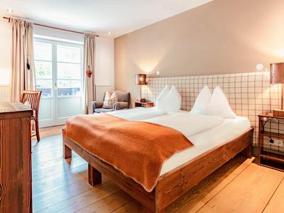 Útulný hotelový pokoj s manželskou postelí, dřevěným nábytkem a balkonem při denním světle.