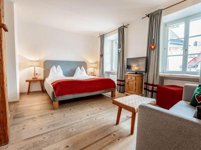 Světý hotelový pokoj s manželskou postelí, dřevěnou podlahou, pohovkou a okny se šedými záclonami.