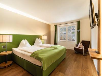 Gemütliches Hotelzimmer mit Doppelbett, grüner Bettdecke, Stuhl und Fenster mit Vorhängen.