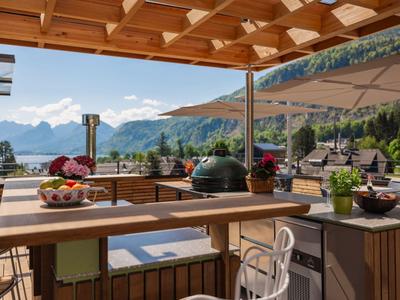 Terrasse mit Essbereich und Grill mit Blick auf See und Berge unter blauem Himmel.