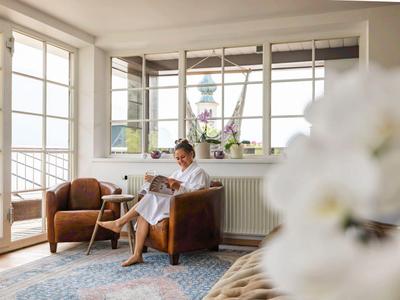 Eine Frau sitzt in einem hellen Zimmer auf einem braunen Sessel und liest ein Buch.