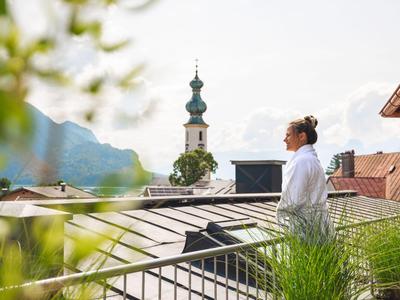 Frau steht auf einem Balkon mit Blick auf Berge und Kirchturm bei klarem Himmel.