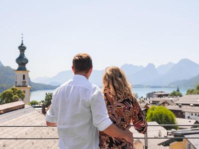 Ein Paar blickt von einem Balkon auf eine Stadt mit Bergen und See im Hintergrund.