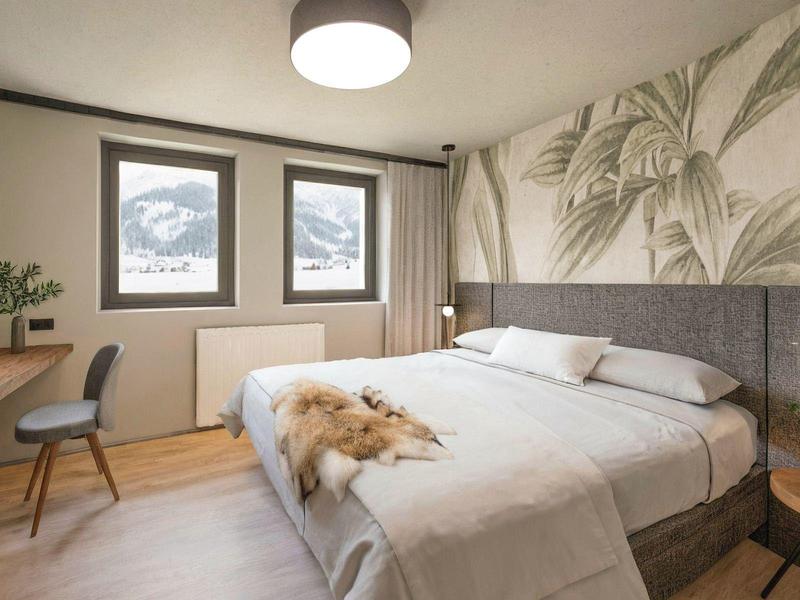 Moderně zařízený hotelový pokoj s dvojlůžkem, židlí, dvěma okny a rostlinami jako dekorací.