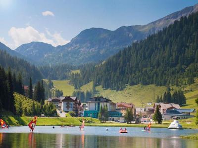 Bergdorf mit See, umgeben von bewaldeten Bergen und Wiesen unter blauem Himmel.