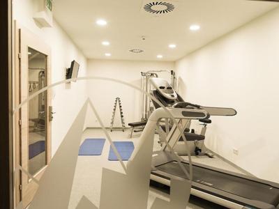 Hell beleuchteter, schmaler Fitnessraum mit Laufband, Hanteln und Übungsmatten.