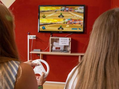 Zwei Personen spielen ein Rennspiel auf einer Videospielkonsole vor einer roten Wand.