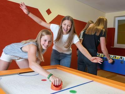 Zwei Mädchen spielen Luft-Hockey, während zwei weitere Personen im Hintergrund bei Tischfußball stehen.