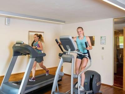 Zwei Frauen trainieren in einem gut beleuchteten Fitnessraum an einem Laufband und einem Crosstrainer.