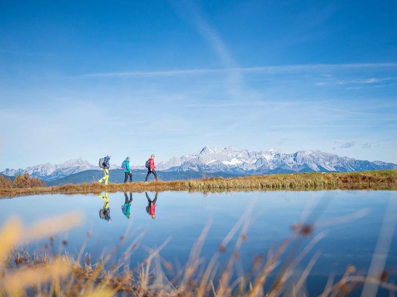 Drei Wanderer gehen an einem See mit Bergkulisse und klarem Himmel entlang.