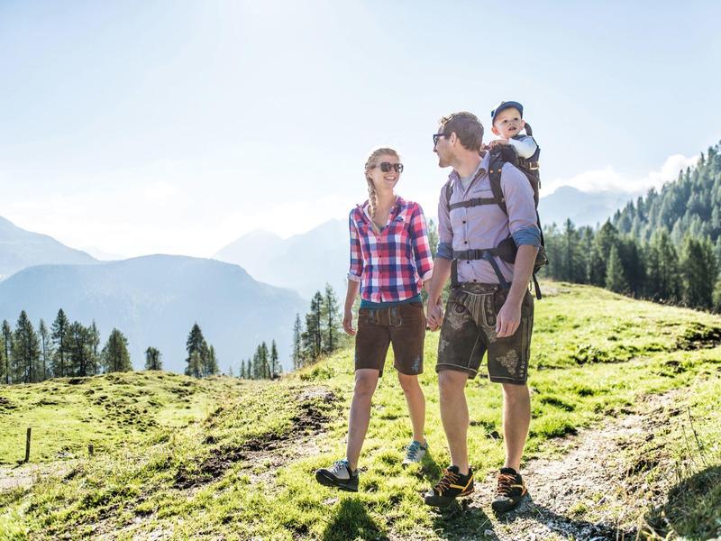 Familie mit Baby wandert auf einem sonnigen Bergweg mit Tannen und Bergen im Hintergrund.