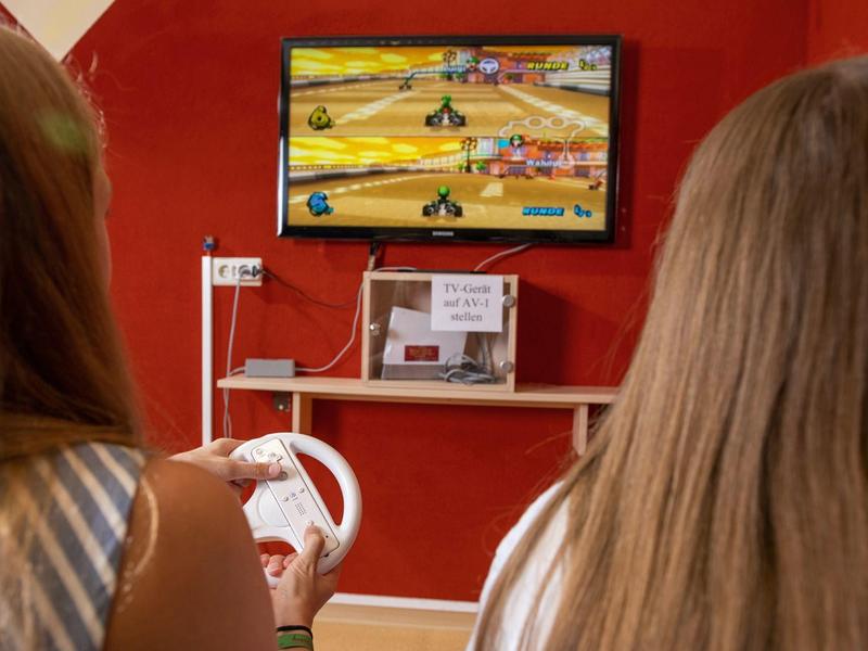 Zwei Personen spielen ein Rennspiel auf einer Videospielkonsole vor einer roten Wand.